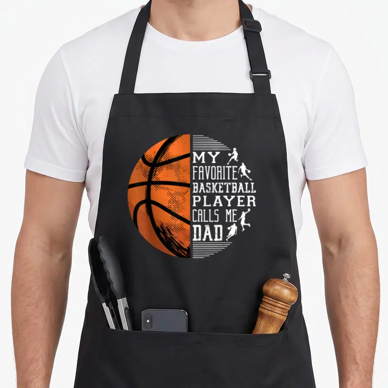 Basketball proud dad apron - Orange half basketball next to stacked white text reading My Favorite Basketball Player — shown on a black cotton canvas apron against a white background