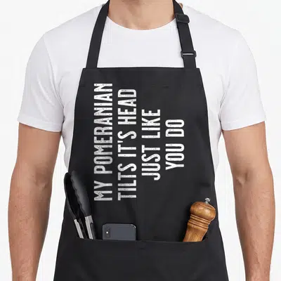 Pomeranian tilt apron - White vertical text reading MY POMERANIAN TILTS IT HEAD JUST LIKE YOU DO — shown on a black cotton canvas apron against a white background