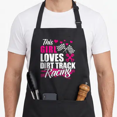 Racer track apron - This GIRL LOVES DIRT TRACK Racing text with pink hearts, checkered flags, and pistons — shown on a black cotton canvas apron against a white background