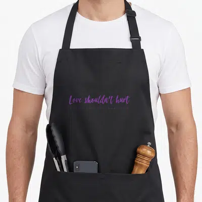 Hurt domestic aprons - Purple text reading Love Shouldn't Hurt and Domestic Violence Awareness stacked vertically — shown on a black cotton canvas apron against a white background
