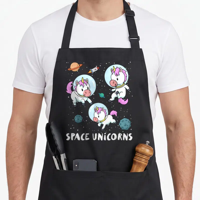 Space unicorn apron - Three cartoon unicorns in helmets floating among planets and stars with text — shown on a black cotton canvas apron against a white background