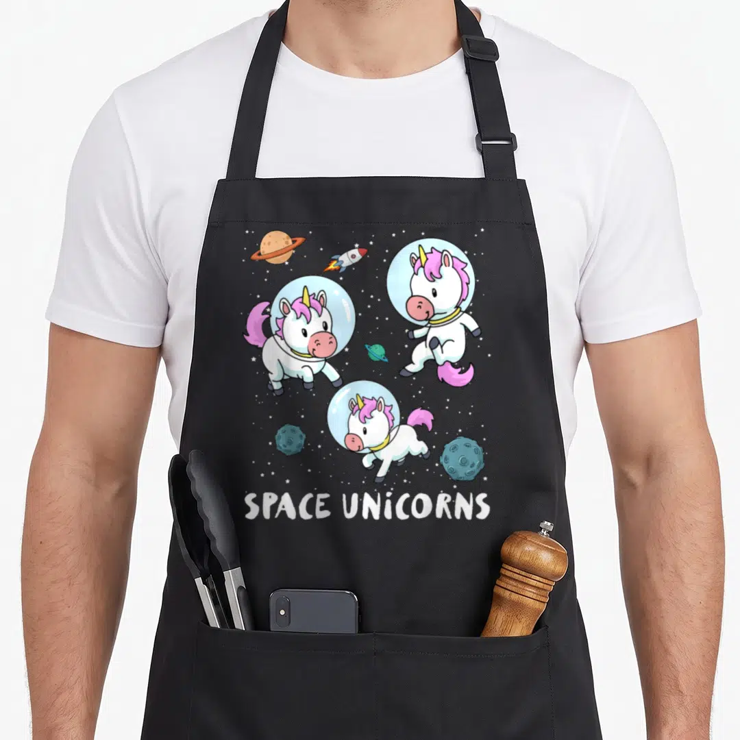 Space unicorn apron - Three cartoon unicorns in helmets floating among planets and stars with text — shown on a black cotton canvas apron against a white background