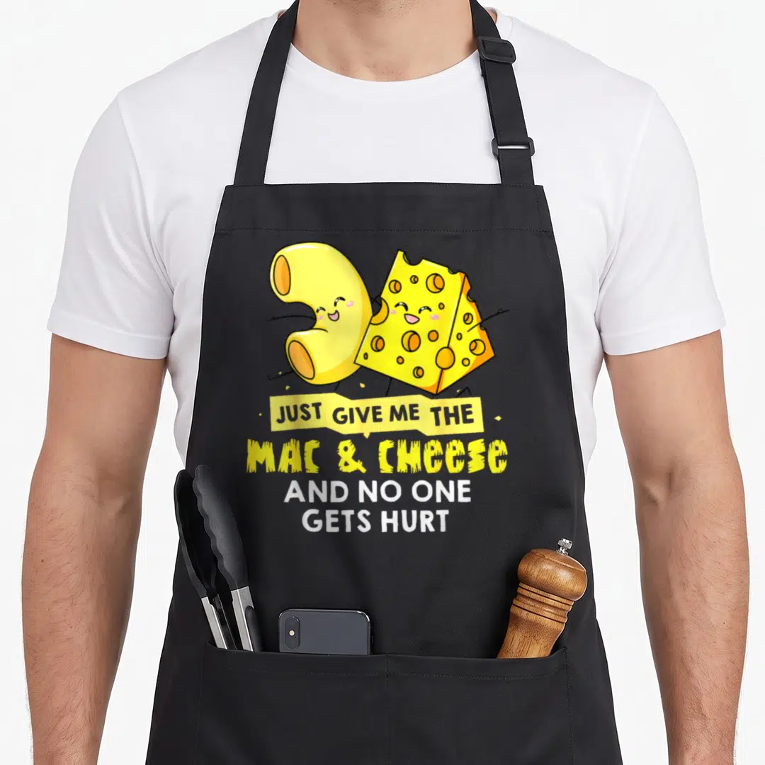 Cheesy mac apron - Cartoon macaroni and cheese character with yellow and white text reading JUST GIVE ME THE MAC & — shown on a black cotton canvas apron against a white background