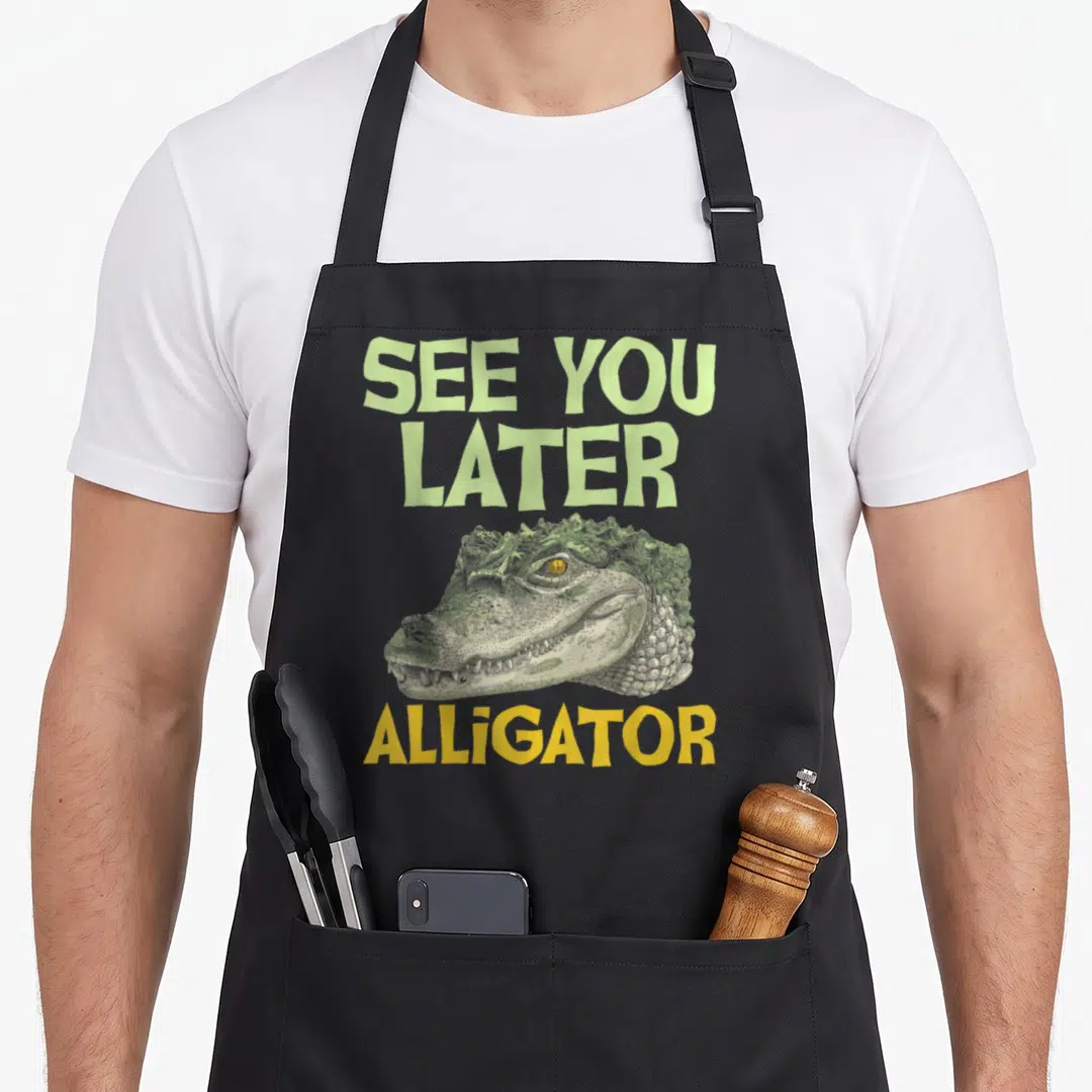 See later animal apron - Cartoon alligator head with SEE YOU LATER ALLIGATOR text in green and yellow — shown on a black cotton canvas apron against a white background