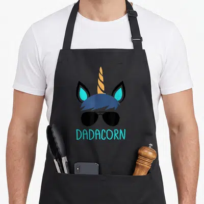 Dadacorn father day apron - Stacked unicorn head with sunglasses and cyan DADACORN text below — shown on a black cotton canvas apron against a white background