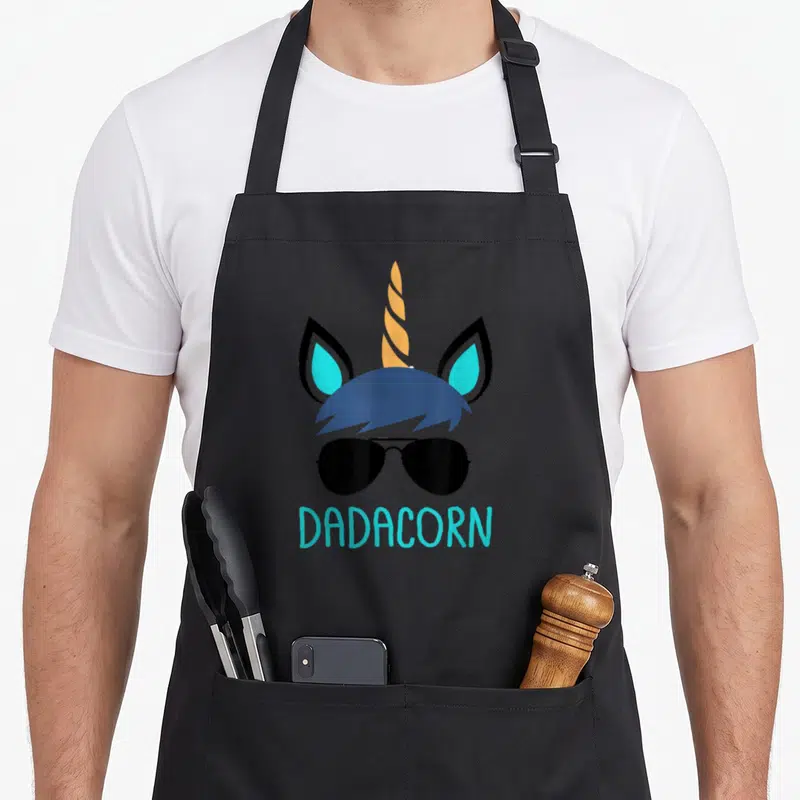 Dadacorn father day apron - Stacked unicorn head with sunglasses and cyan DADACORN text below — shown on a black cotton canvas apron against a white background