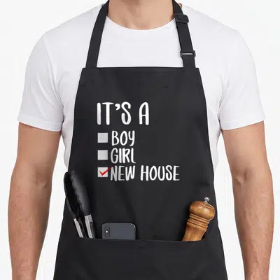 Cool occasion novelty apron - White checklist text IT'S A BOY GIRL NEW HOUSE with red checkmark — shown on a black cotton canvas apron against a white background