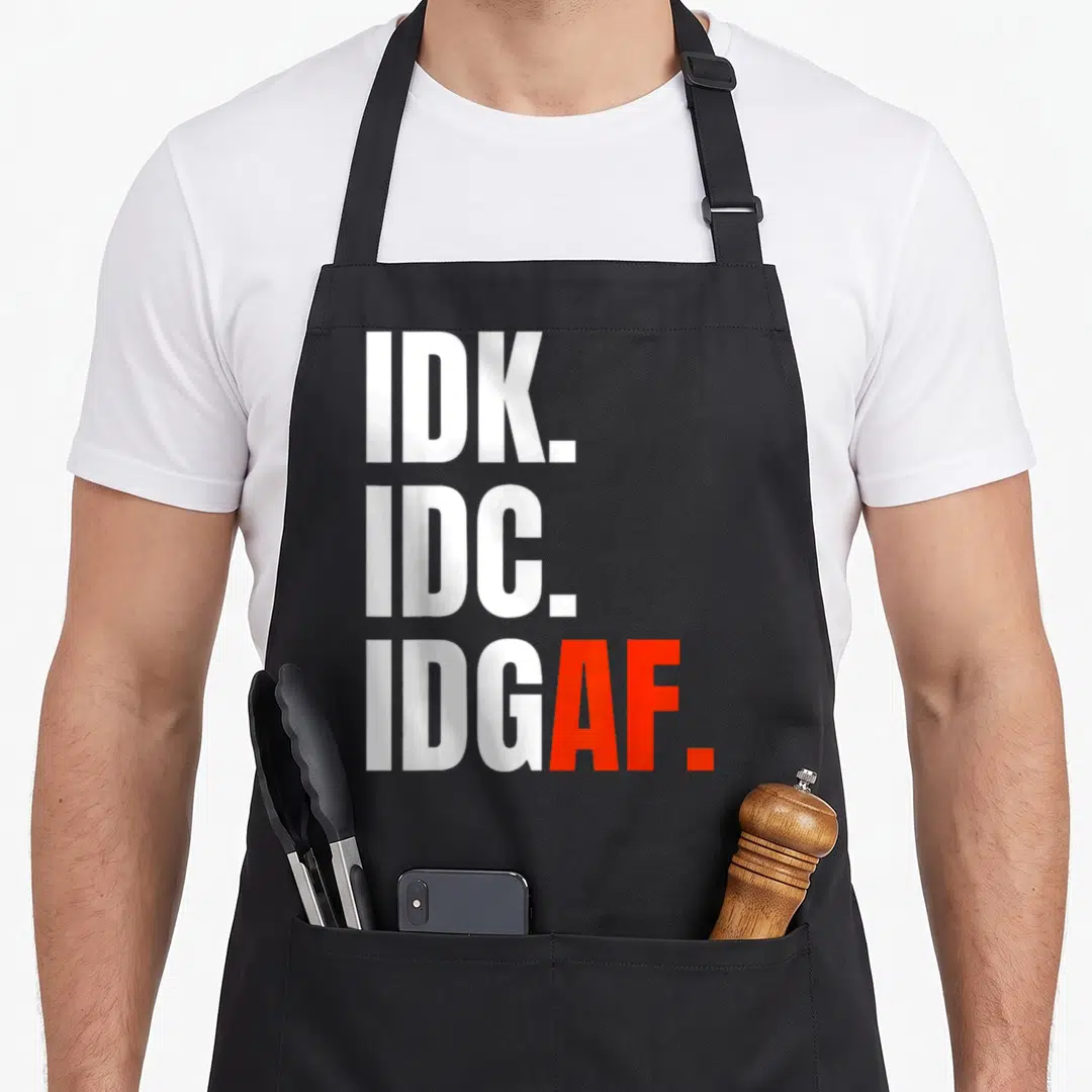 Funny idk idgaf apron - Stacked white IDK IDC IDGAF text with red AF and period on gray background — shown on a black cotton canvas apron against a white background