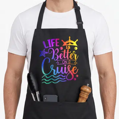 Better family aprons - Gradient text Life is Better on a Cruise with ship wheel, starfish, waves, and bubbles — shown on a black cotton canvas apron against a white background