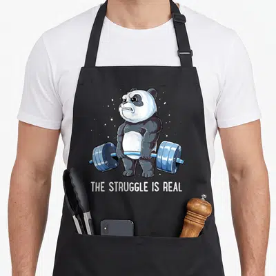 Fitness deadlift apron - Muscular cartoon panda lifting blue barbell with white dots and text THE STRUGGLE IS REAL — shown on a black cotton canvas apron against a white background