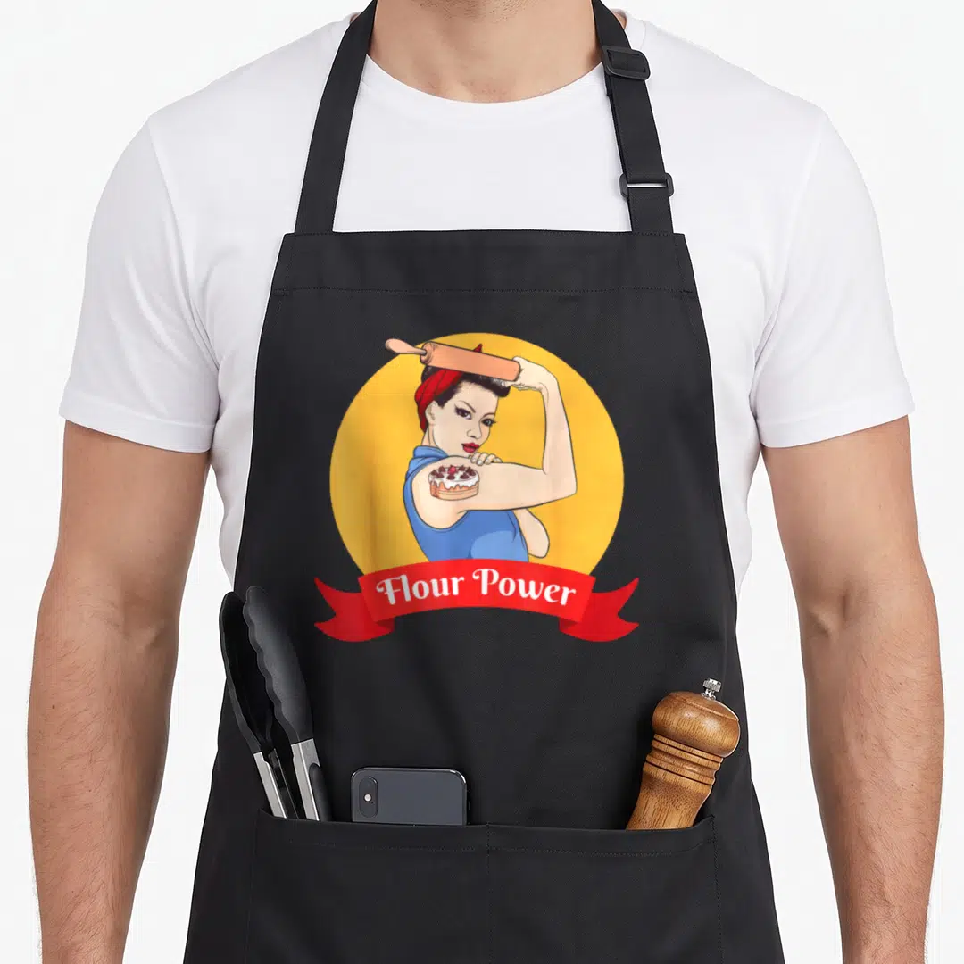 Pastry baker apron - Retro woman flexing cake on bicep with rolling pin on shoulder inside yellow circle with Flour — shown on a black cotton canvas apron against a white background