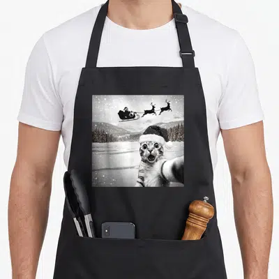Santa funny cat apron - Black and white photo of a cat wearing a Santa hat taking a selfie with Santa's sleigh — shown on a black cotton canvas apron against a white background