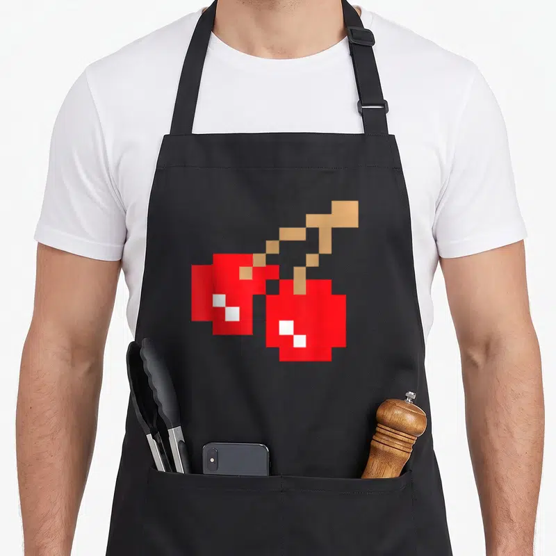 Arcade cherry apron - Two red pixelated cherries with tan stems and white highlights in 8-bit art style — shown on a black cotton canvas apron against a white background