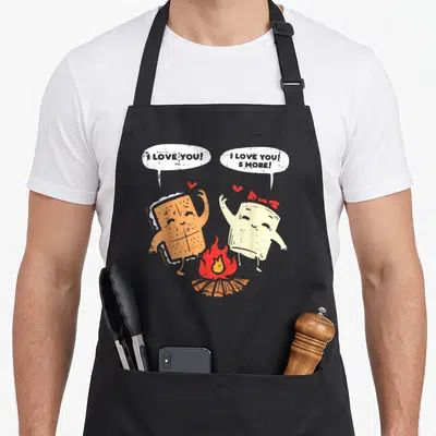 Smore couple apron - Two cartoon s'mores by campfire with I Love You S'More speech bubbles and red hearts — shown on a black cotton canvas apron against a white background