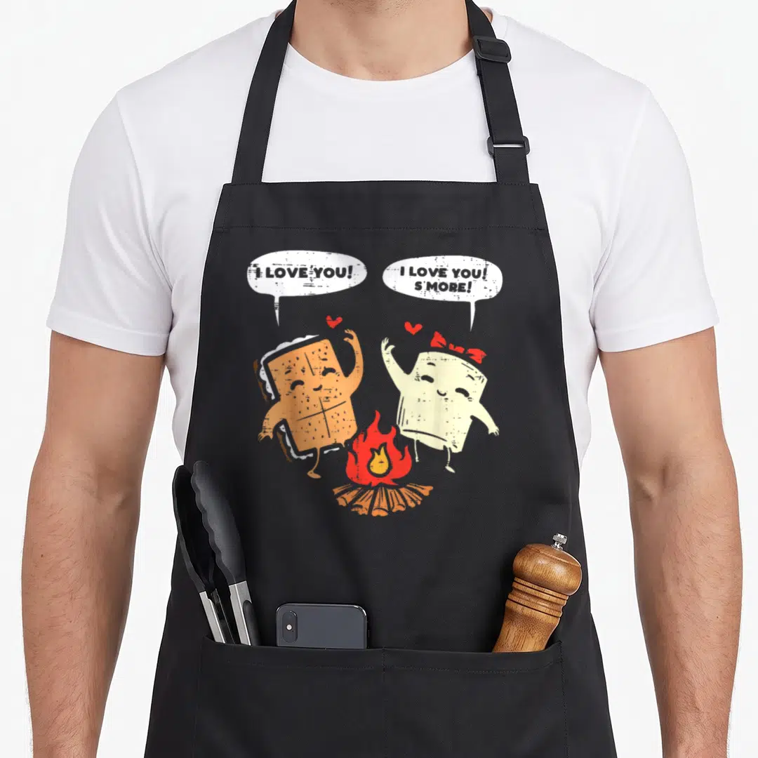 Smore couple apron - Two cartoon s'mores by campfire with I Love You S'More speech bubbles and red hearts — shown on a black cotton canvas apron against a white background
