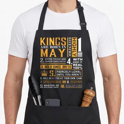 May king apron - Orange and white typography listing ten sarcastic personality traits for May birthdays on a grey — shown on a black cotton canvas apron against a white background