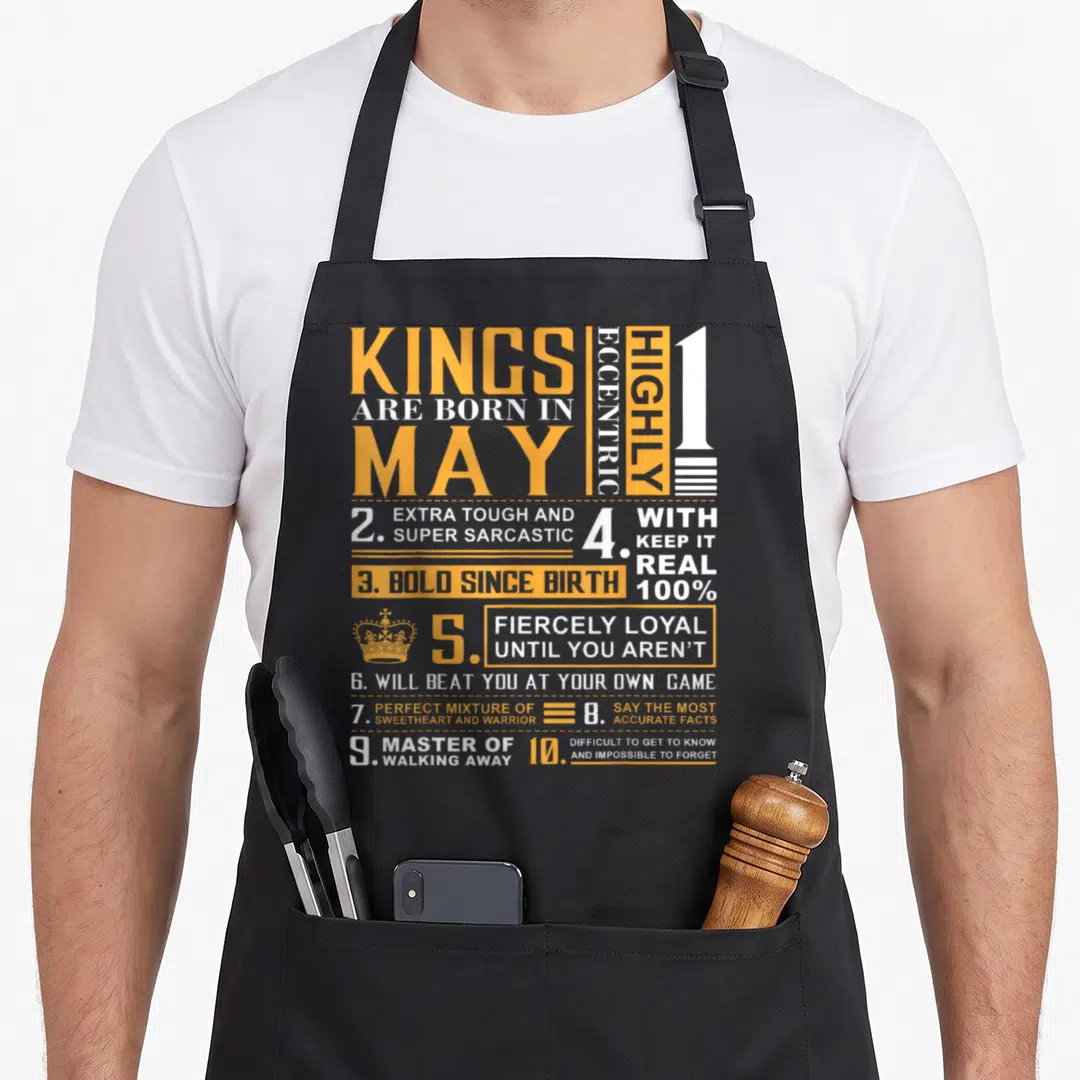 May king apron - Orange and white typography listing ten sarcastic personality traits for May birthdays on a grey — shown on a black cotton canvas apron against a white background