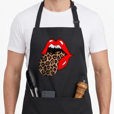 Lip leopard print apron - Red lips and white teeth with a leopard print tongue hanging down to the left — shown on a black cotton canvas apron against a white background