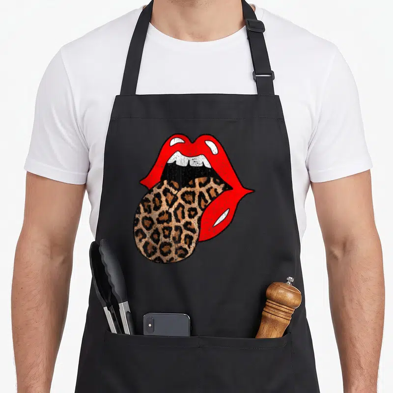 Lip leopard print apron - Red lips and white teeth with a leopard print tongue hanging down to the left — shown on a black cotton canvas apron against a white background