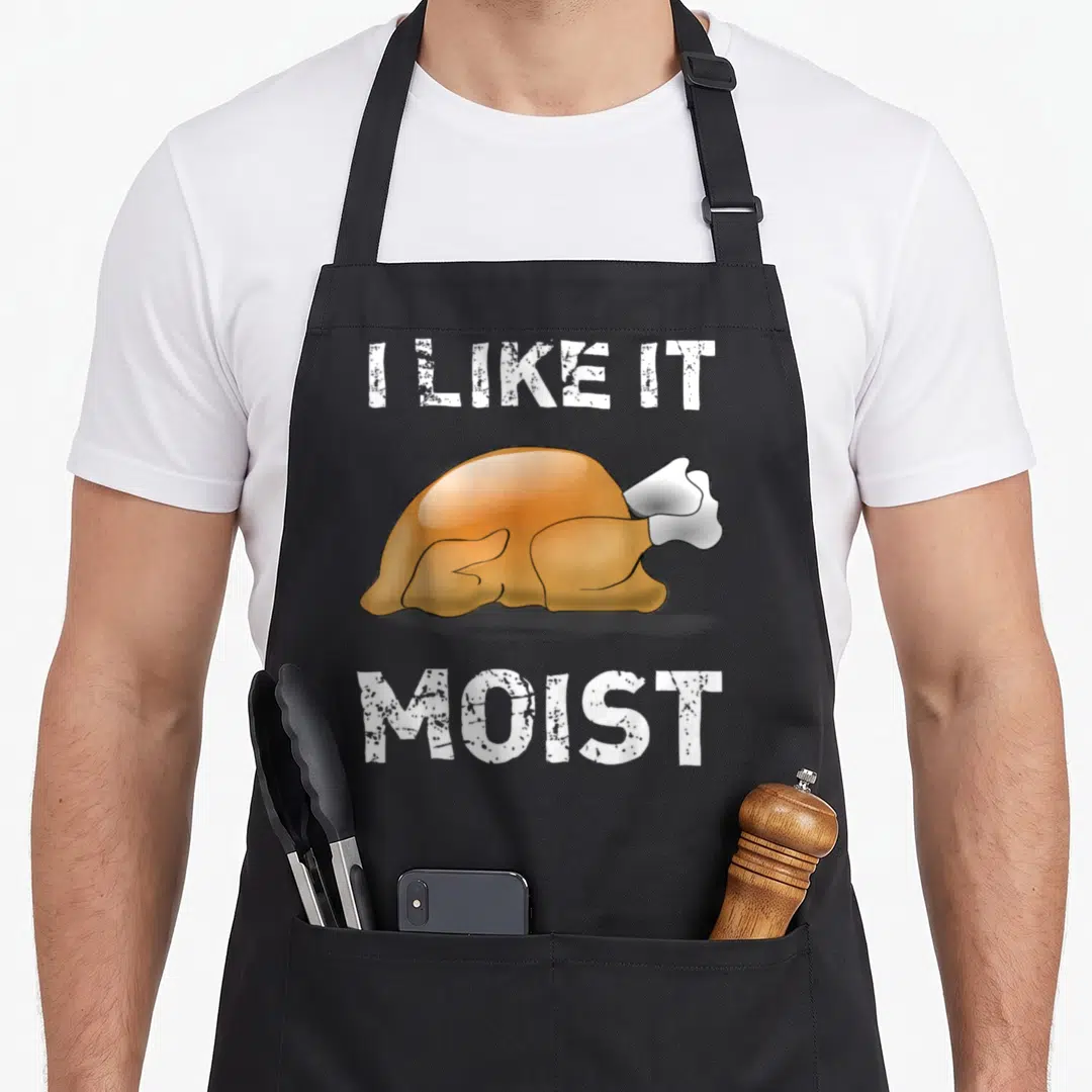 Moist turkey thanksgiving apron - Cartoon roasted turkey between I LIKE IT MOIST text — shown on a black cotton canvas apron against a white background