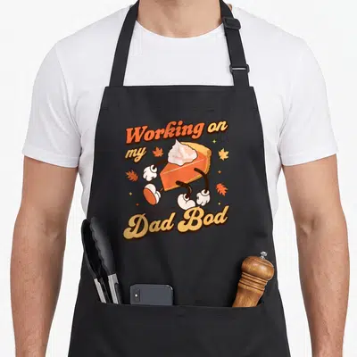 Father thanksgiving apron - Cartoon pie slice with arms and legs and text Working on my Dad Bod surrounded by leaves — shown on a black cotton canvas apron against a white background