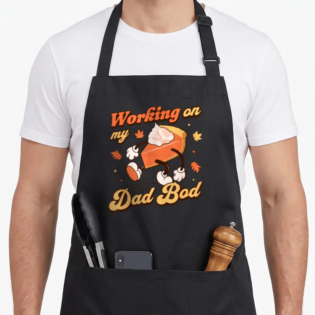 Father thanksgiving apron - Cartoon pie slice with arms and legs and text Working on my Dad Bod surrounded by leaves — shown on a black cotton canvas apron against a white background