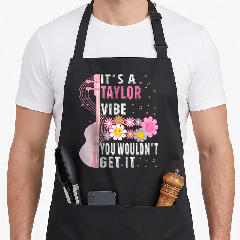 Taylor groovy apron - Pink guitar, smiling flowers, and text IT'S A TAYLOR VIBE YOU WOULDN'T GET IT — shown on a black cotton canvas apron against a white background