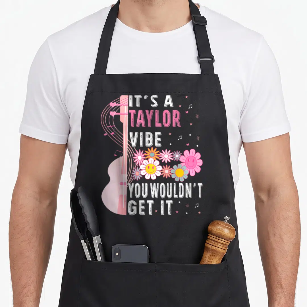 Taylor groovy apron - Pink guitar, smiling flowers, and text IT'S A TAYLOR VIBE YOU WOULDN'T GET IT — shown on a black cotton canvas apron against a white background