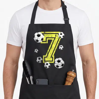Fan birthday apron - Yellow number seven with six distressed soccer balls in black and white — shown on a black cotton canvas apron against a white background