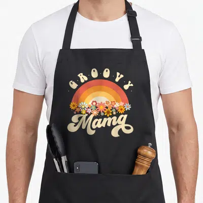 Groovy colorful aprons - Retro rainbow with colorful flowers and groovy Mama text in cream, red, orange, and yellow — shown on a black cotton canvas apron against a white background