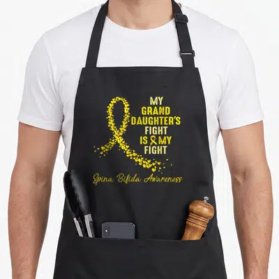 Granddaughter spina bifida awareness apron - Yellow ribbon made of small hearts next to bold text reading MY GRAND — shown on a black cotton canvas apron against a white background