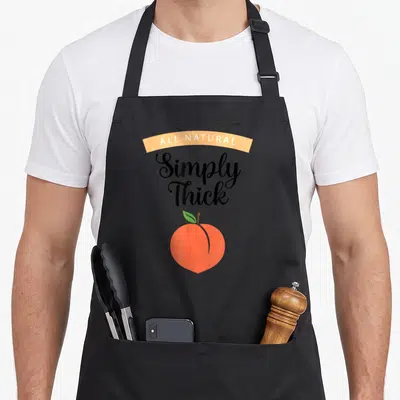 Simply body apron - All Natural Simply Thick text above orange peach with green leaf — shown on a black cotton canvas apron against a white background