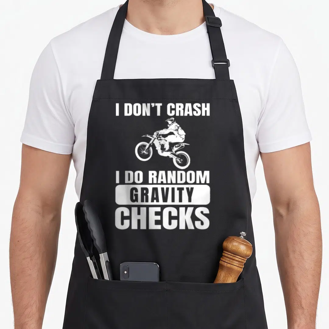 Crash bike apron - I DON'T CRASH I DO RANDOM GRAVITY CHECKS motocross rider white typography — shown on a black cotton canvas apron against a white background