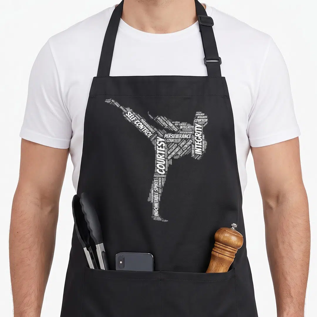 Fighter taekwondo apron - White typography word cloud forming a high kick with Courtesy, Integrity, Perseverance, Self — shown on a black cotton canvas apron against a white background
