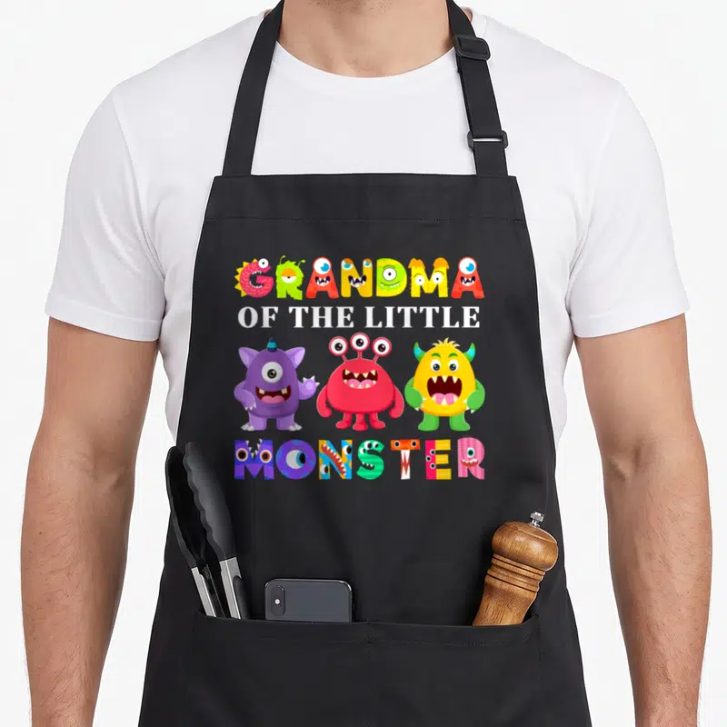 Monster grandma in apron - Three colorful cartoon monsters below GRANDMA OF THE LITTLE MONSTER text in white — shown on a black cotton canvas apron against a white background