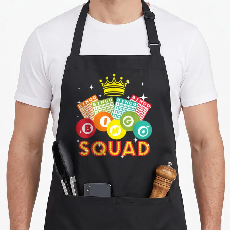 Retro player bingo apron - Yellow crown above four fanned bingo cards and five colored balls spelling BINGO SQUAD — shown on a black cotton canvas apron against a white background