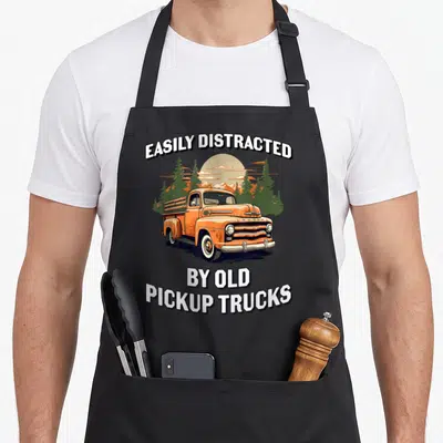 Vintage easily truck apron - Orange cartoon pickup truck with trees and mountains and text easily distracted by old — shown on a black cotton canvas apron against a white background