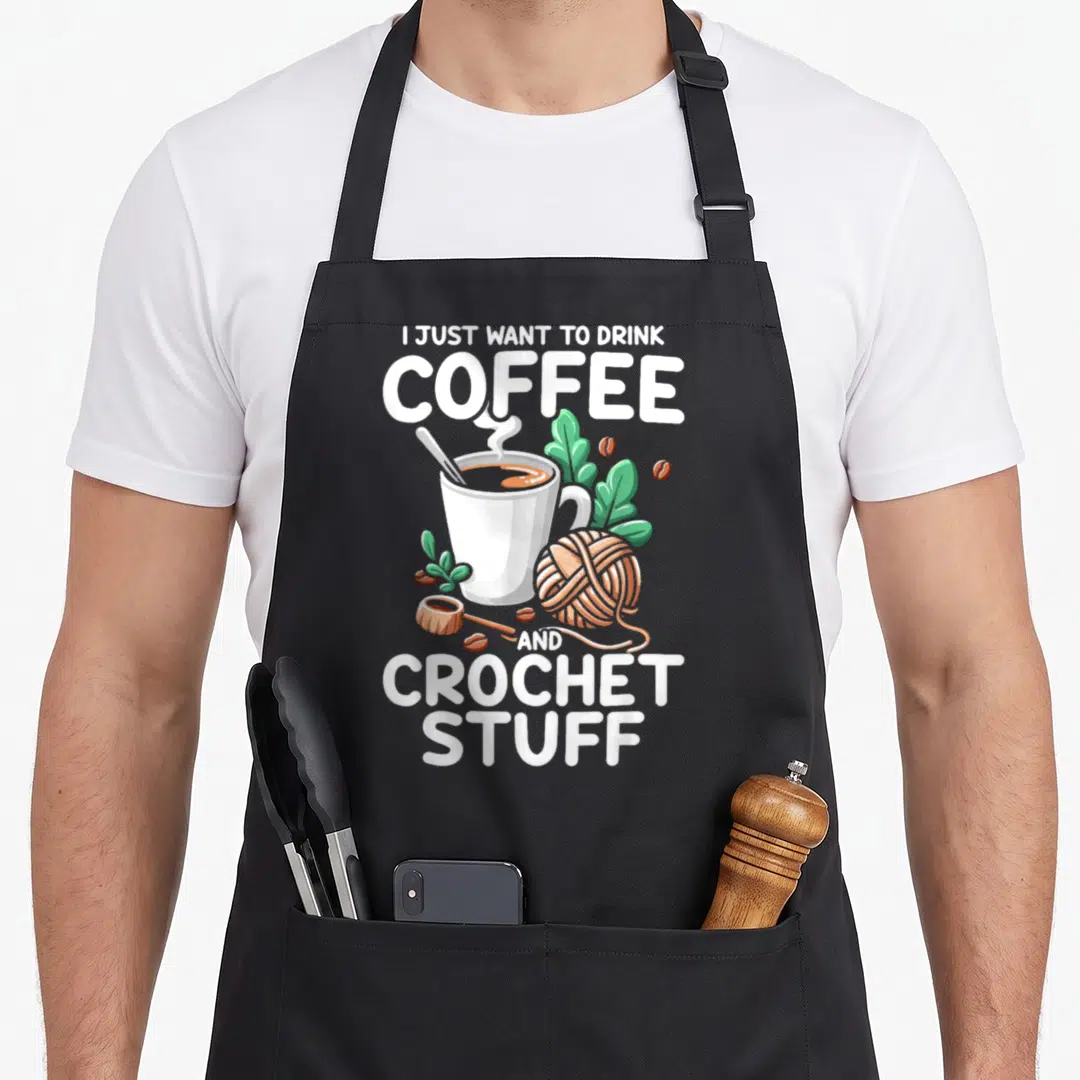 Drink crochet apron - Stacked text reading I JUST WANT TO DRINK COFFEE AND CROCHET STUFF around a cup, yarn, beans — shown on a black cotton canvas apron against a white background