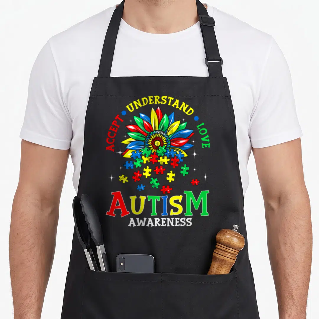 Autism golf mom apron - Multi-colored flower with falling puzzle pieces and Accept Understand Love text — shown on a black cotton canvas apron against a white background