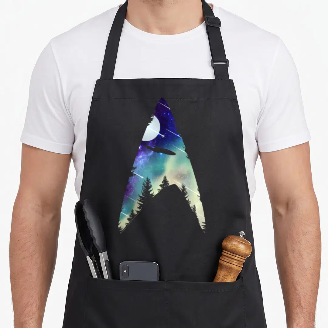 Galactic star apron - Starfleet delta shape containing a moon, spaceship, and pine trees in purple and blue nebula — shown on a black cotton canvas apron against a white background