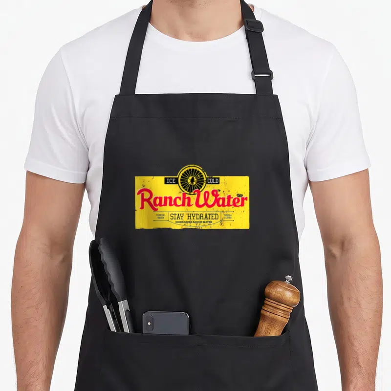 Drink southern aprons - Yellow rectangle with black wagon wheel, red Ranch Water script, and teal text listing tequila — shown on a black cotton canvas apron against a white background