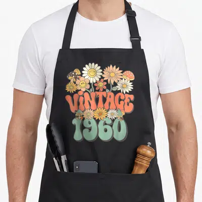 Hippie floral aprons - Vintage 1960 text with orange and mint green cartoon flowers and mushrooms — shown on a black cotton canvas apron against a white background
