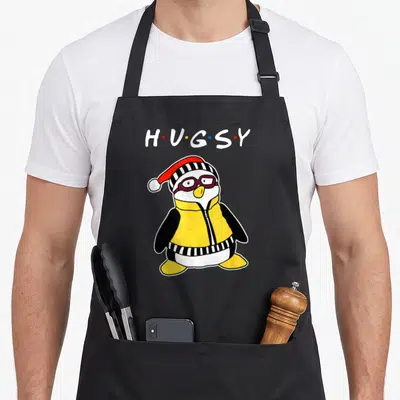 Hugsy penguin aprons - HUGSY text above cartoon penguin in yellow vest and striped hat — shown on a black cotton canvas apron against a white background