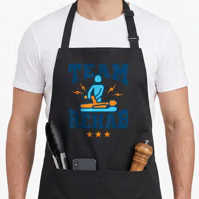Rehab my therapy apron - Arched Team Rehab text surrounding a blue therapist and orange patient with lightning bolts — shown on a black cotton canvas apron against a white background