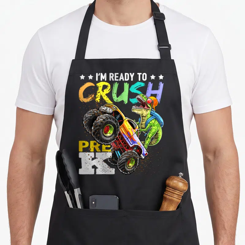 School truck apron - Cartoon T-Rex with backpack beside monster truck, colorful CRUSH PRE K text with mud splatters — shown on a black cotton canvas apron against a white background