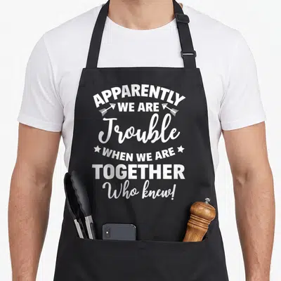 Apparently friends who apron - White stacked text with arrows and stars reading apparently we are trouble when we are — shown on a black cotton canvas apron against a white background