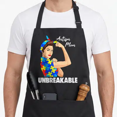Awareness golf mom apron - Cartoon woman flexing arm in puzzle shirt with Autism Mom and UNBREAKABLE text — shown on a black cotton canvas apron against a white background