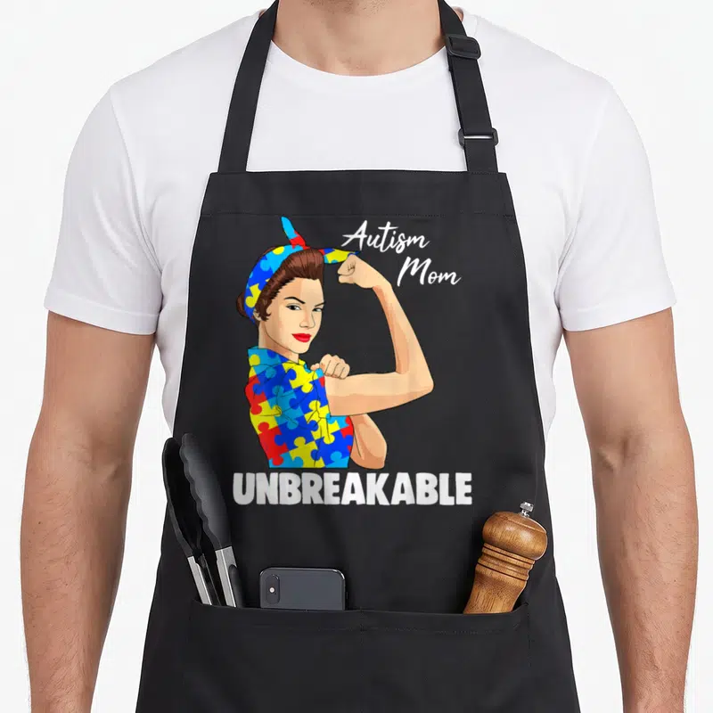 Awareness golf mom apron - Cartoon woman flexing arm in puzzle shirt with Autism Mom and UNBREAKABLE text — shown on a black cotton canvas apron against a white background