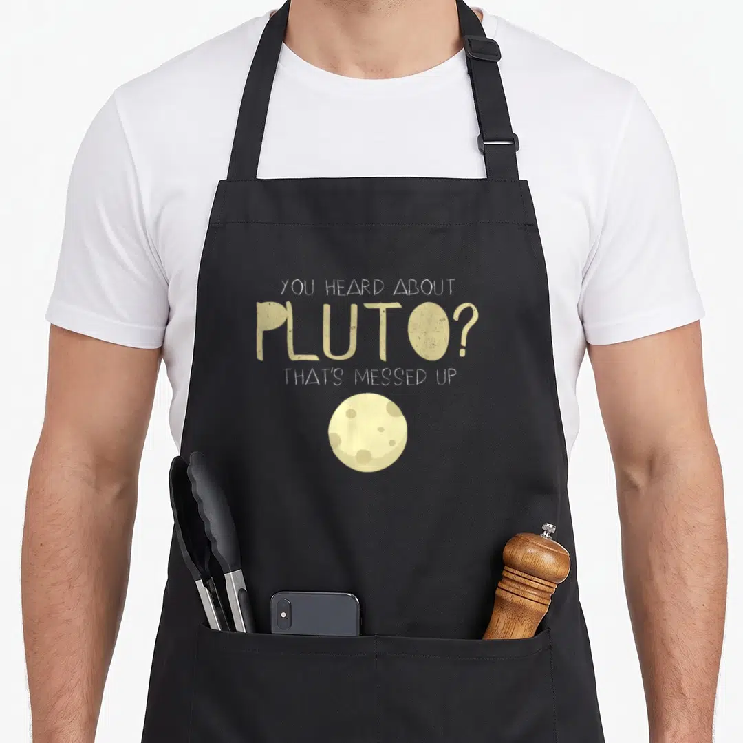Pluto novelty apron - Beige text You Heard About Pluto That's Messed Up above small circular planet illustration — shown on a black cotton canvas apron against a white background