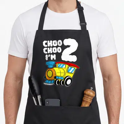 Train 2nd apron - Choo Choo I'm 2 cloud text above yellow blue red cartoon train — shown on a black cotton canvas apron against a white background